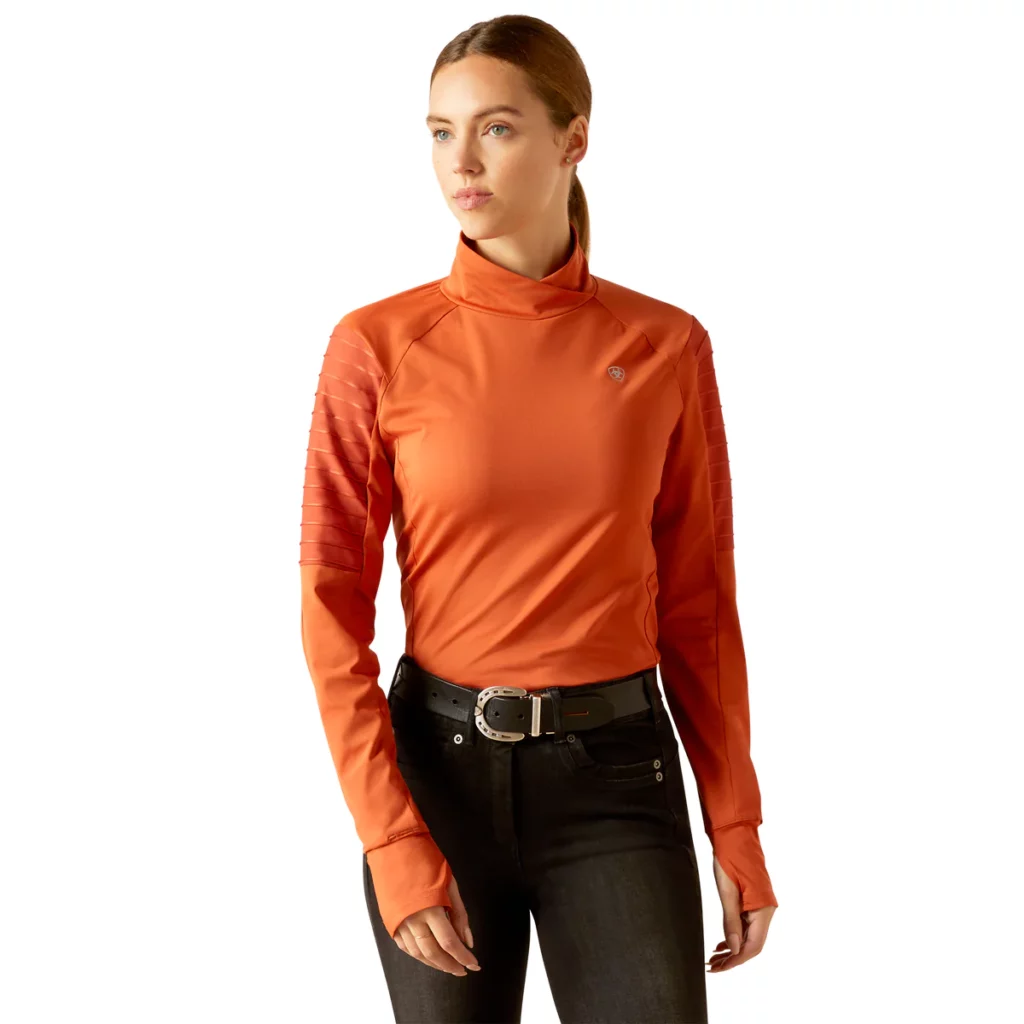 Woman wearing an orange Ariat long sleeve baselayer with a tall collar